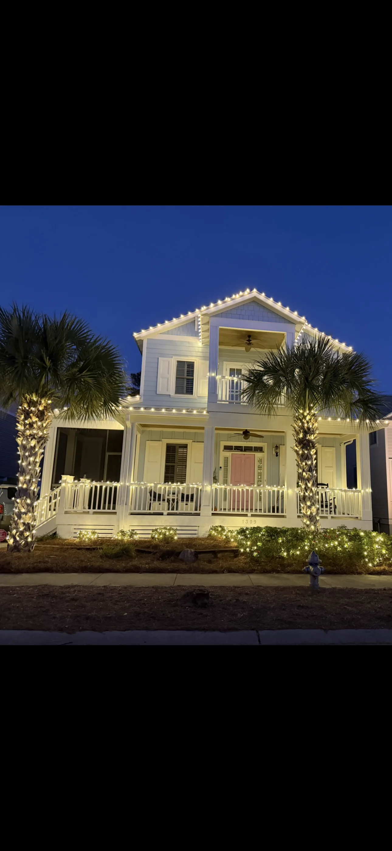 White two-story home with wrapped porch, pressure-washed siding, white railings, and landscape lighting on palm trees