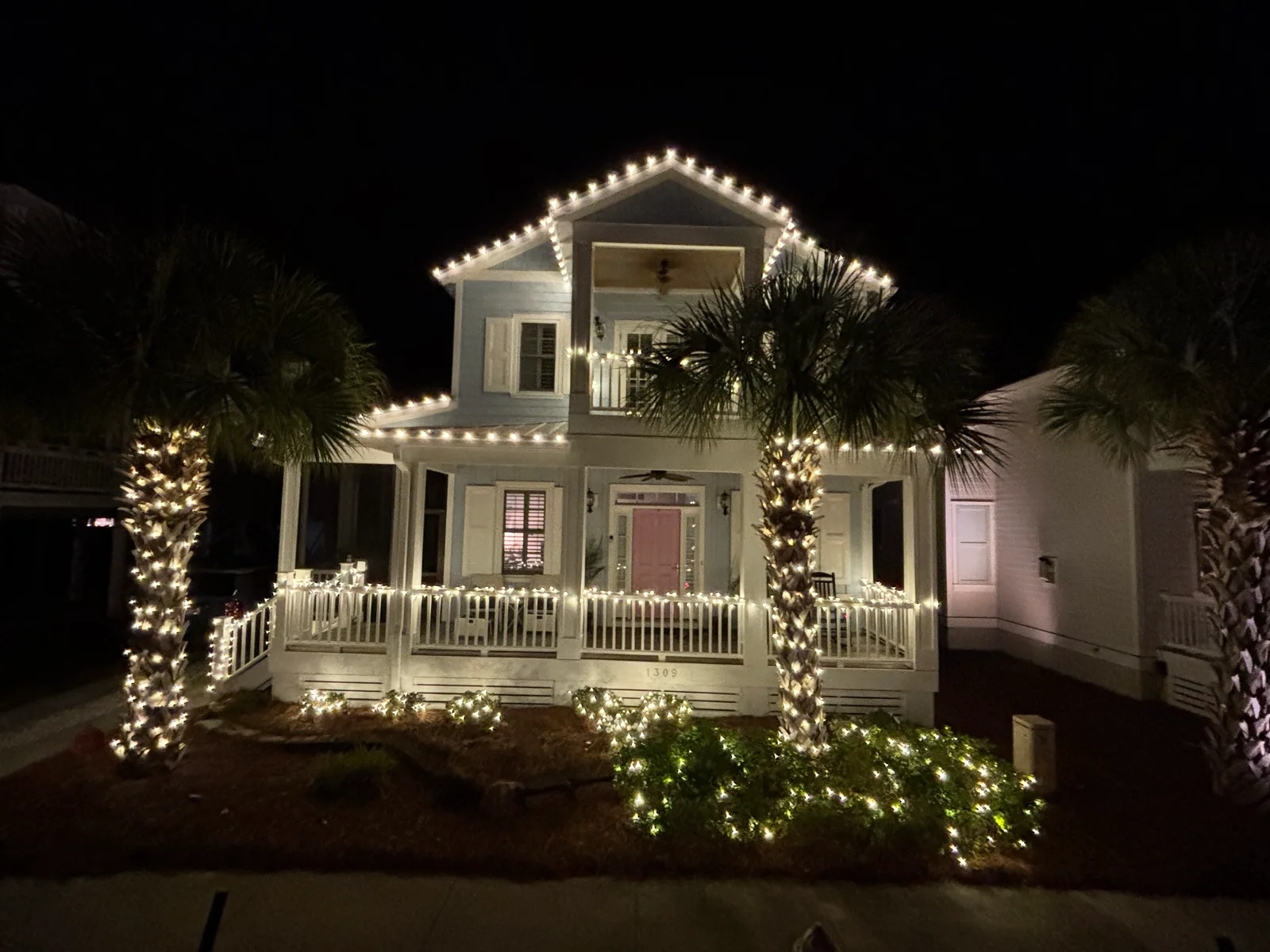 Two-story white home with LED string lights on roofline, palm trees, and porch railings at night