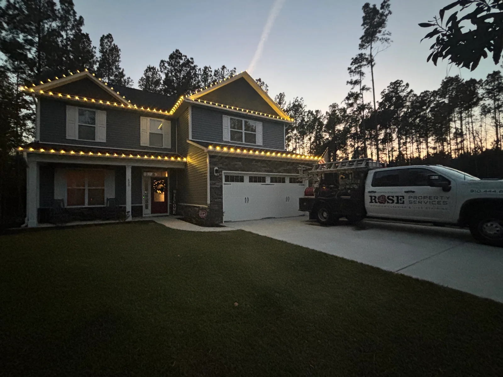 Two-story home with warm white string lights installed along roofline and gable peaks at dusk